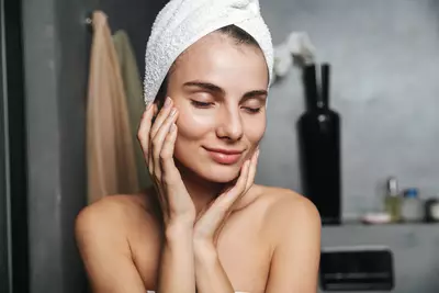 Mulher com toalha branca enrolada no cabelo apos aplicar hidratante de rosto