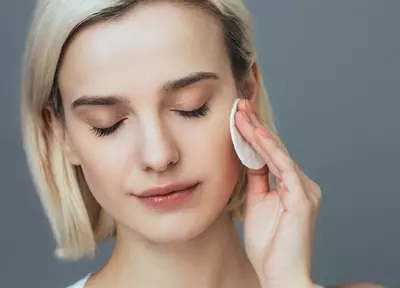 Jovem mulher loira aplicando tonico facial e agua micelar no rosto com algodao