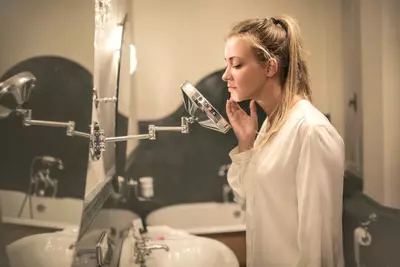 Mulher adulta aplicando produto para tratamento da acne da mulher adulta em frente ao espelho, com produtos de skincare ao fundo.