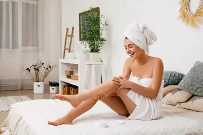 Mulher branca enrolada em uma toalha apos o banho sentada em sua cama aplicando hidratante corporal em sua perna.