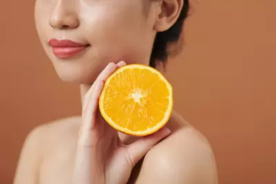 Foto de mulher segurando fatia de laranja proxima ao rosto simbolizando beneficios da vitamina C para a pele
