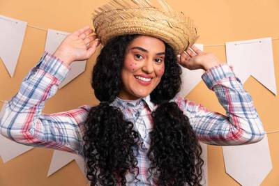 Mulher sorrindo com chapeu de palha, maquiagem de sardas e cabelo encaracolado, com penteado de festa junina.