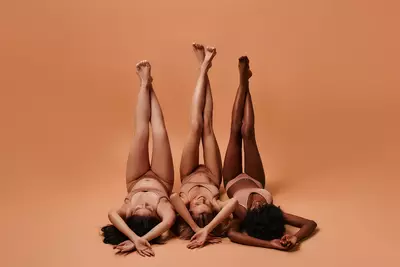 Tres mulheres deitadas lado a lado no chao utilizando roupas intimas bege com as pernas cruzadas para cima e os bracos cruzados para cima da cabeca, mostrando como a pele fica lisa e sem marcas apos o clareamento intimo.