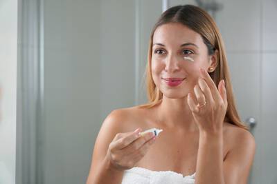 Mulher sorrindo, aplicando creme clareador proximo ao olho em frente ao espelho do banheiro, segurando um pequeno tubo de produto na outra mao.