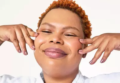 Mulher sorridente com pele hidratada e luminosa, usando roupao branco, toca suavemente o rosto apos a rotina de skincare, exemplo de pele bem cuidada.