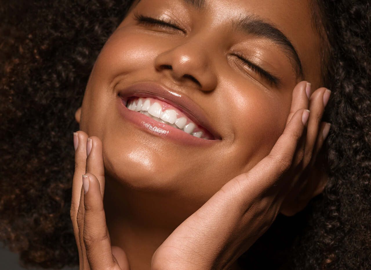Mulher jovem sorrindo com os olhos fechados e as maos no rosto, exibindo pele hidratada e radiante apos a rotina de skincare com hidratante facial.