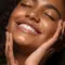 Mulher jovem sorrindo com os olhos fechados e as maos no rosto, exibindo pele hidratada e radiante apos a rotina de skincare com hidratante facial.