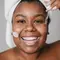 Mulher negra com cabelos presos em uma toalha aplicando um serum anti-idade transparente no rosto.