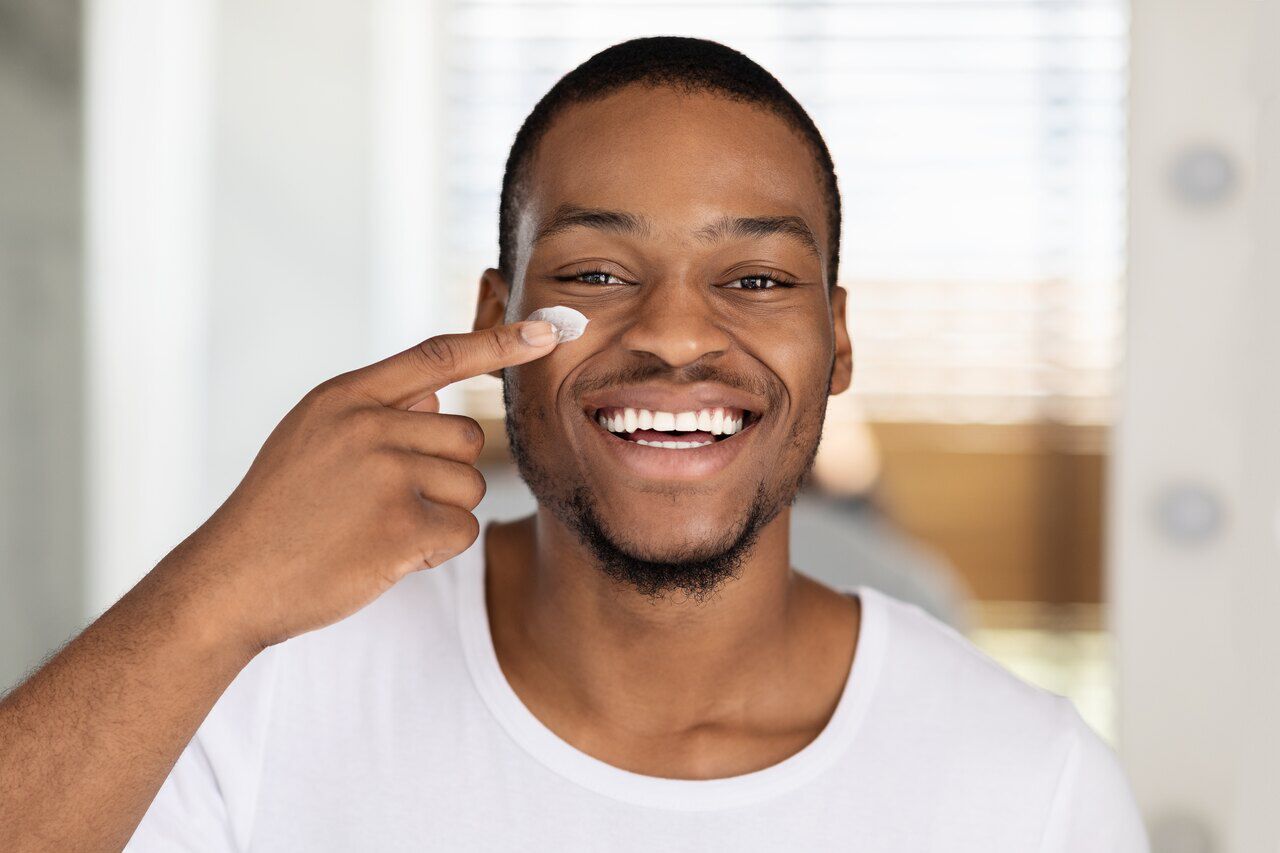 Homem jovem negro sorrindo e  passando hidratante masculino no rosto