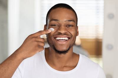 Homem jovem negro sorrindo e  passando hidratante masculino no rosto