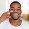 Homem jovem negro sorrindo e passando hidratante masculino no rosto