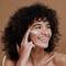 Mulher com cabelos encaracolados e pele morena sorrindo enquanto aplica protetor solar facial branco na bochecha direita, olhando para o lado.