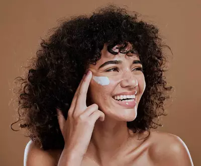 Mulher com cabelos encaracolados e pele morena sorrindo enquanto aplica protetor solar facial branco na bochecha direita, olhando para o lado.