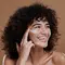 Mulher com cabelos encaracolados e pele morena sorrindo enquanto aplica protetor solar facial branco na bochecha direita, olhando para o lado.