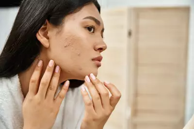 I Mulher sorridente aplicando produto no rosto em frente ao espelho. Ao fundo, uma prateleira com diversos produtos de skincare e maquiagem.