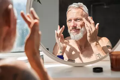 Homem grisalho com barba branca aplica creme no rosto enquanto se olha no espelho, em um ambiente iluminado pela luz natural. Ele aparece sem camisa e possui tatuagens nos bracos.