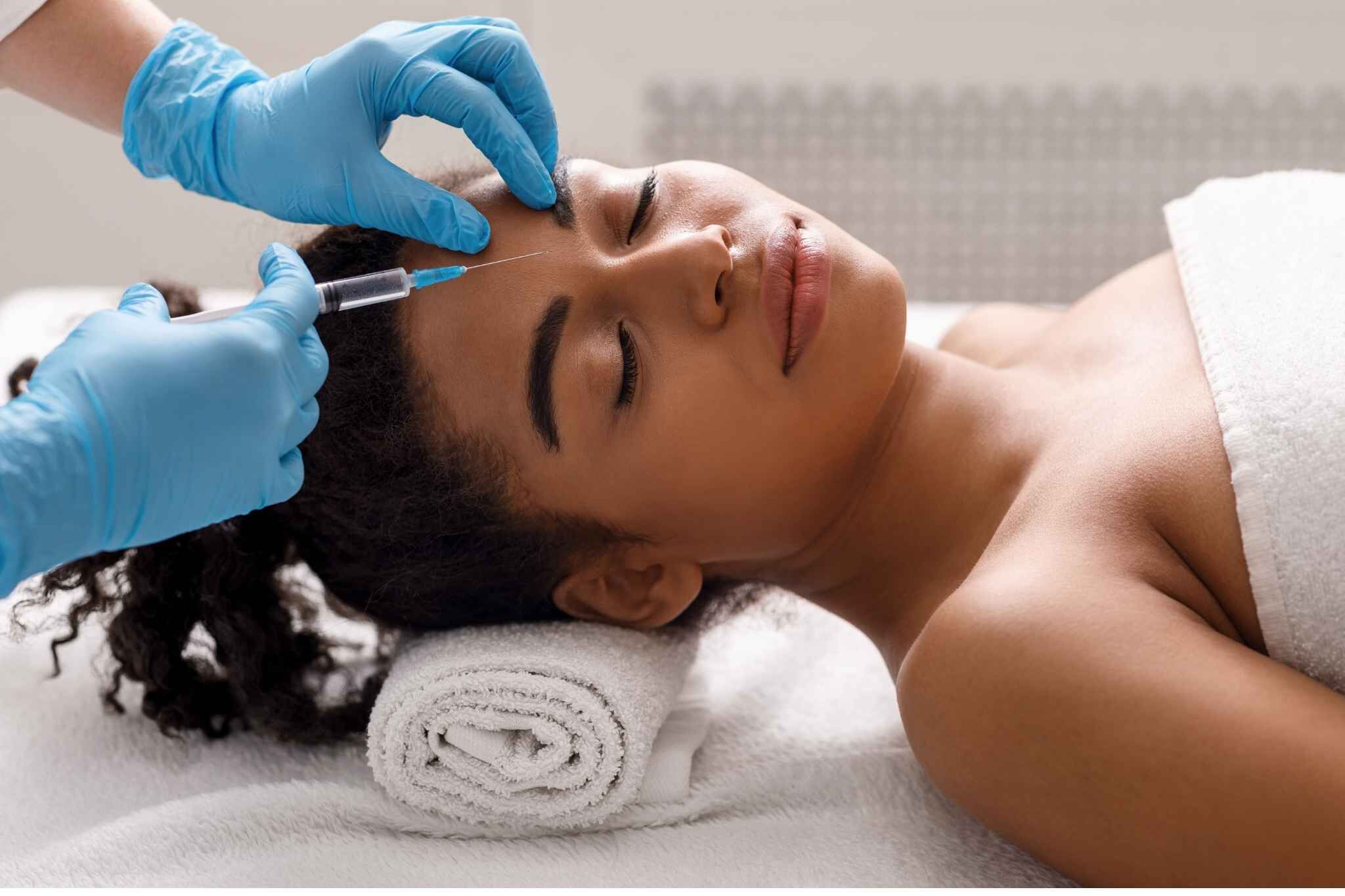 Mulher deitada em maca recebendo aplicacao de injecao facial feita por profissional com luvas azuis, em ambiente de clinica dermatologica.