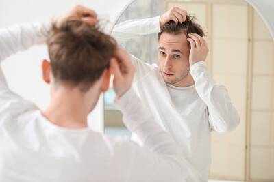 Homem branco se olhando no espelho observando o couro cabeludo em busca de diminuicao da quantidade de fios e queda de cabelo