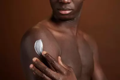Homem negro sem camisa aplicando hidratante corporal com ceramidas no braco com as maos.