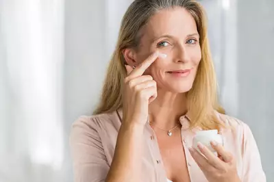 Mulher de 50 anos aplicando creme para a area dos olhos com o dedo indicador.