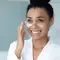 Mulher negra aplicando creme para olheira embaixo dos olhos com o dedo indicador enquanto sorri.