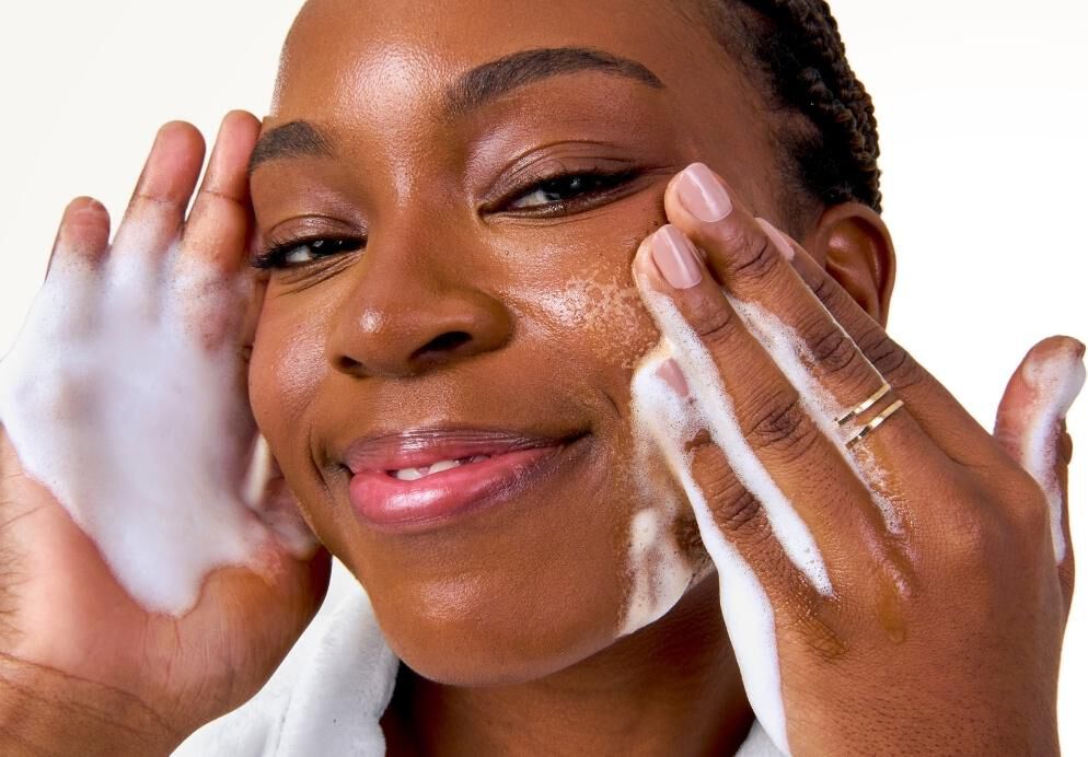 Mulher sorridente de roupao branco aplicando espuma de limpeza facial com movimentos suaves, ilustrando a etapa de limpeza na rotina de skincare.