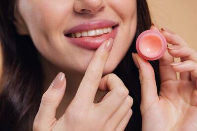 Mulher sorridente aplicando hidratante labial rosado de um pequeno pote, com labios bem cuidados e unhas com esmalte nude em fundo bege neutro.