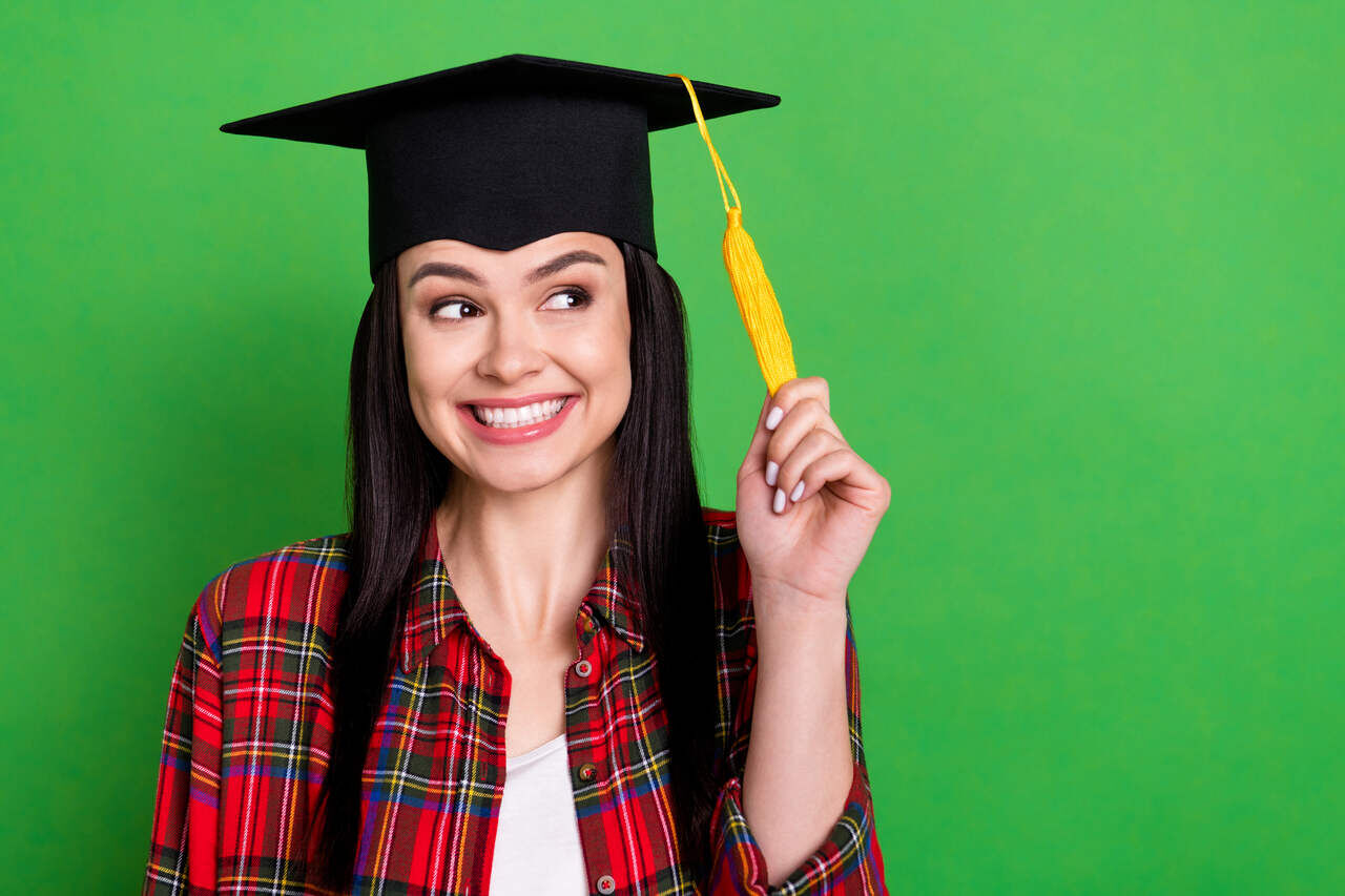 Mulher jovem sorrindo com chapeu de formatura, segurando o tassel amarelo, usando camisa xadrez vermelha sobre fundo verde para representar penteados de formatura
