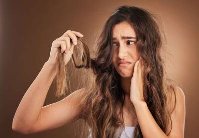 Mulher jovem com expressao preocupada segurando uma mecha de cabelo longo, ressecado e com frizz, observando os fios danificados com frustracao.