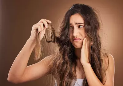 Mulher jovem com expressao preocupada segurando uma mecha de cabelo longo, ressecado e com frizz, observando os fios danificados com frustracao.