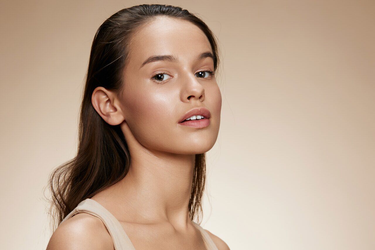 Jovem mulher com pele luminosa e maquiagem natural em tons neutros, cabelo castanho preso para tras, sobre fundo bege claro em estilo editorial.