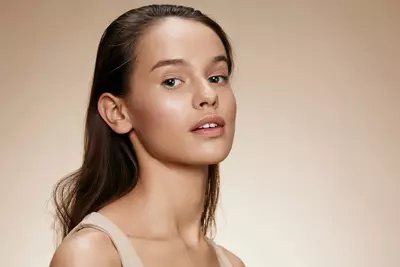 Jovem mulher com pele luminosa e maquiagem natural em tons neutros, cabelo castanho preso para tras, sobre fundo bege claro em estilo editorial.