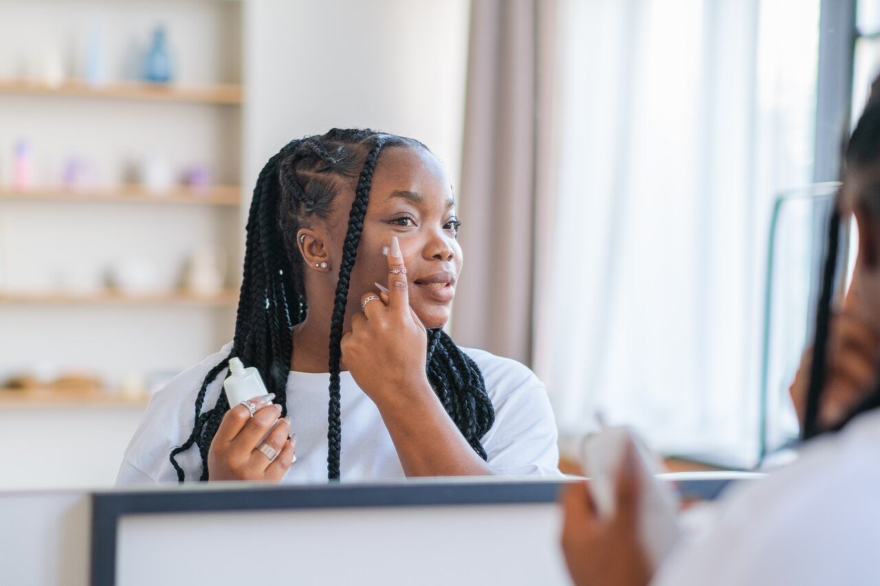 Mulher negra aplicando produto de skincare no rosto enquanto se olha no espelho, representando a importância da rotina de cuidados com a pele durante o uso de canetas emagrecedoras.