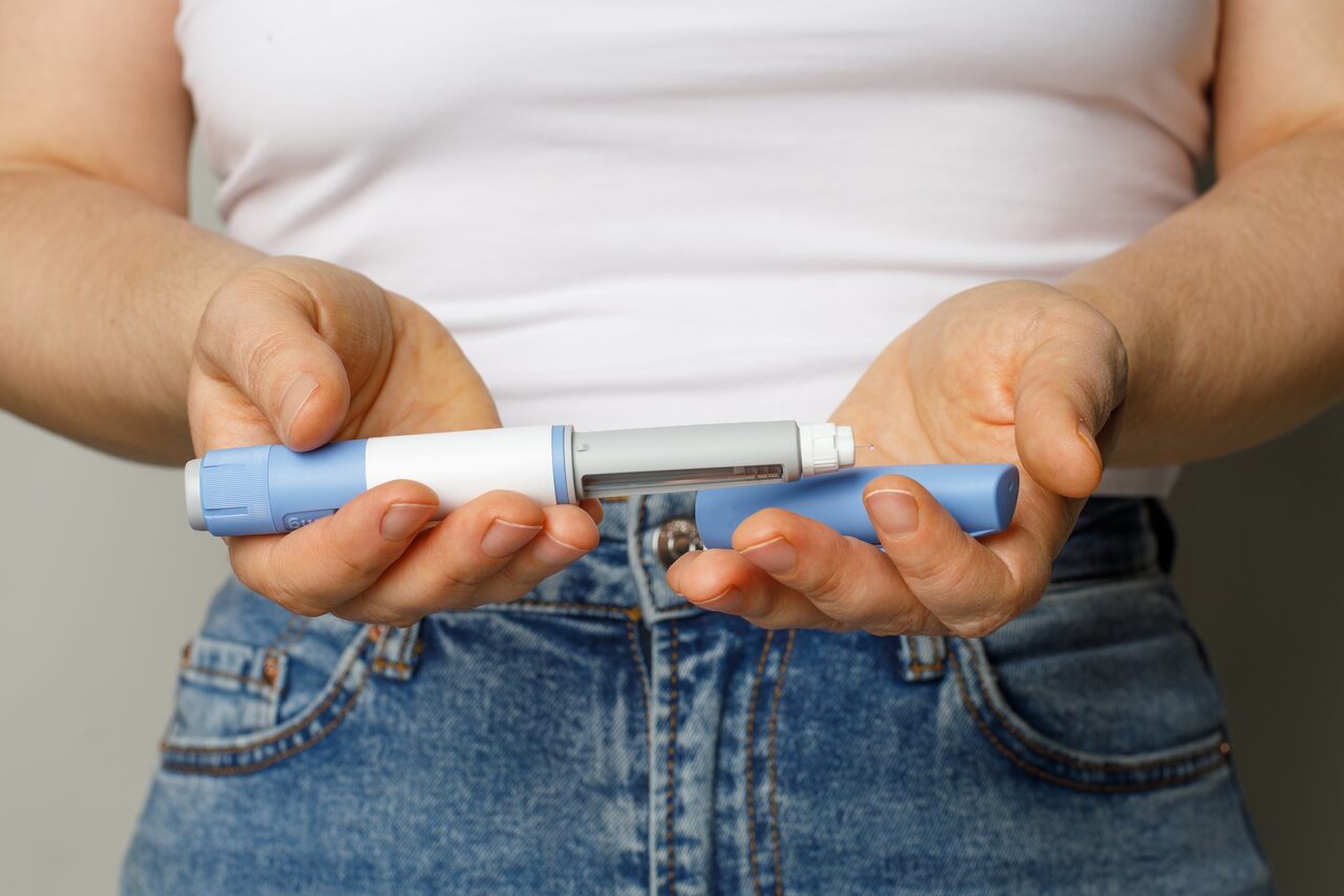 Uma pessoa, vestindo uma camiseta branca e jeans azul, segura uma caneta injetora tipo Ozempic azul e branca horizontalmente com as duas mãos