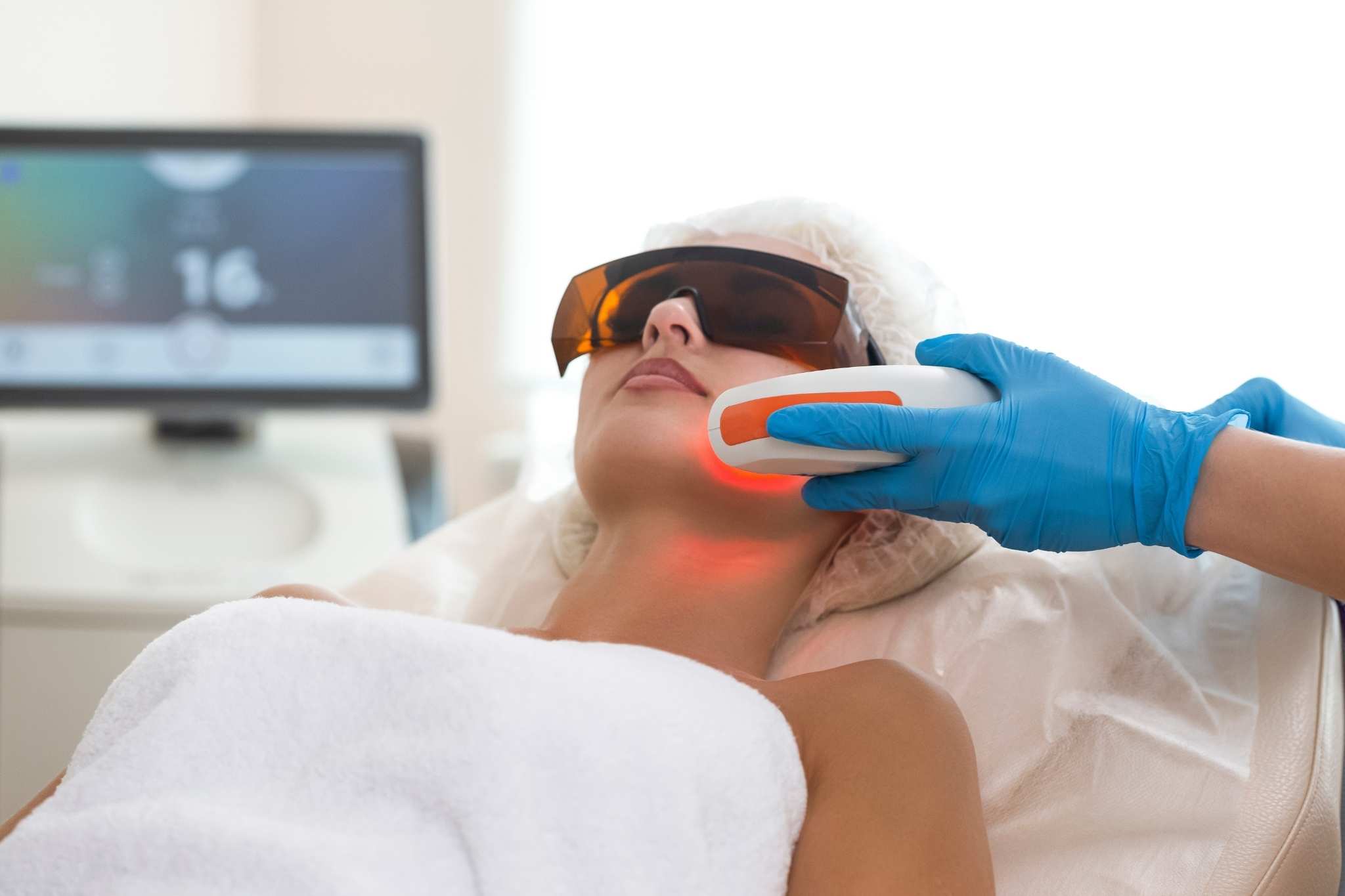 Tratamento com laser facial sendo realizado como preparação para o Projeto Verão durante o inverno, com pessoa usando óculos de proteção e recebendo aplicação de luz por profissional com luvas azuis