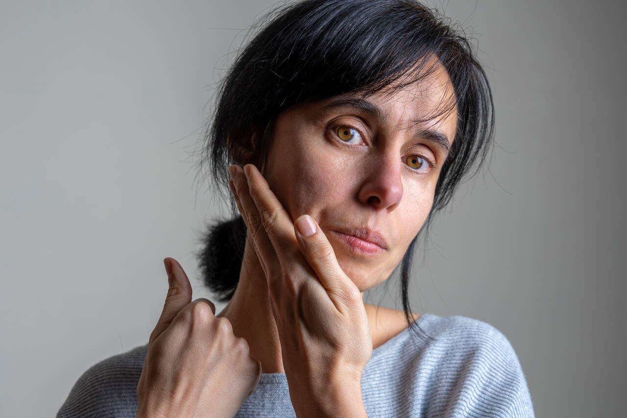 Uma mulher com cabelo escuro preso, olhando diretamente para a câmera enquanto puxa a pele da bochecha e do pescoço com uma mão. 