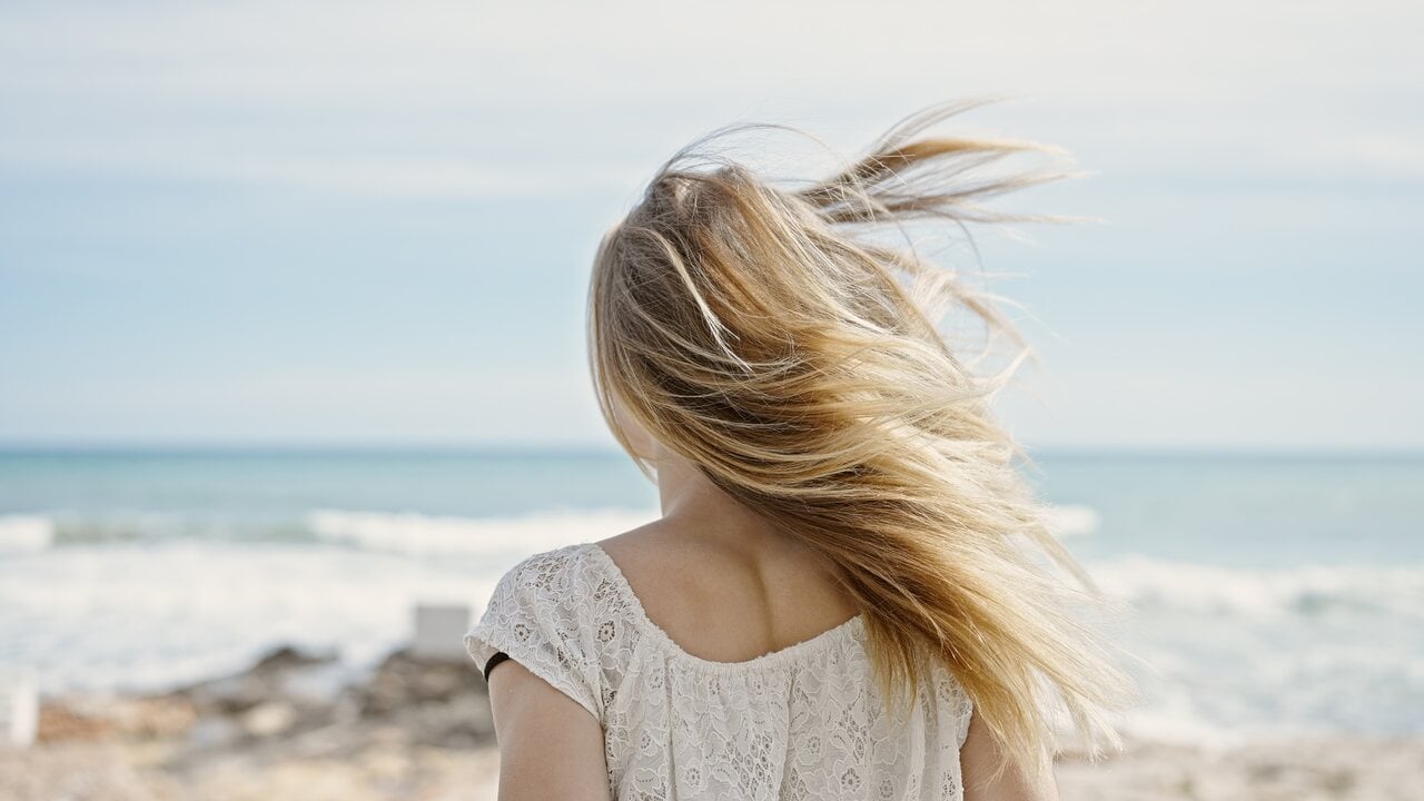 Mulher loira de costas com cabelo ao vento, vestindo blusa rendada clara, em praia com mar e céu azul ao fundo.