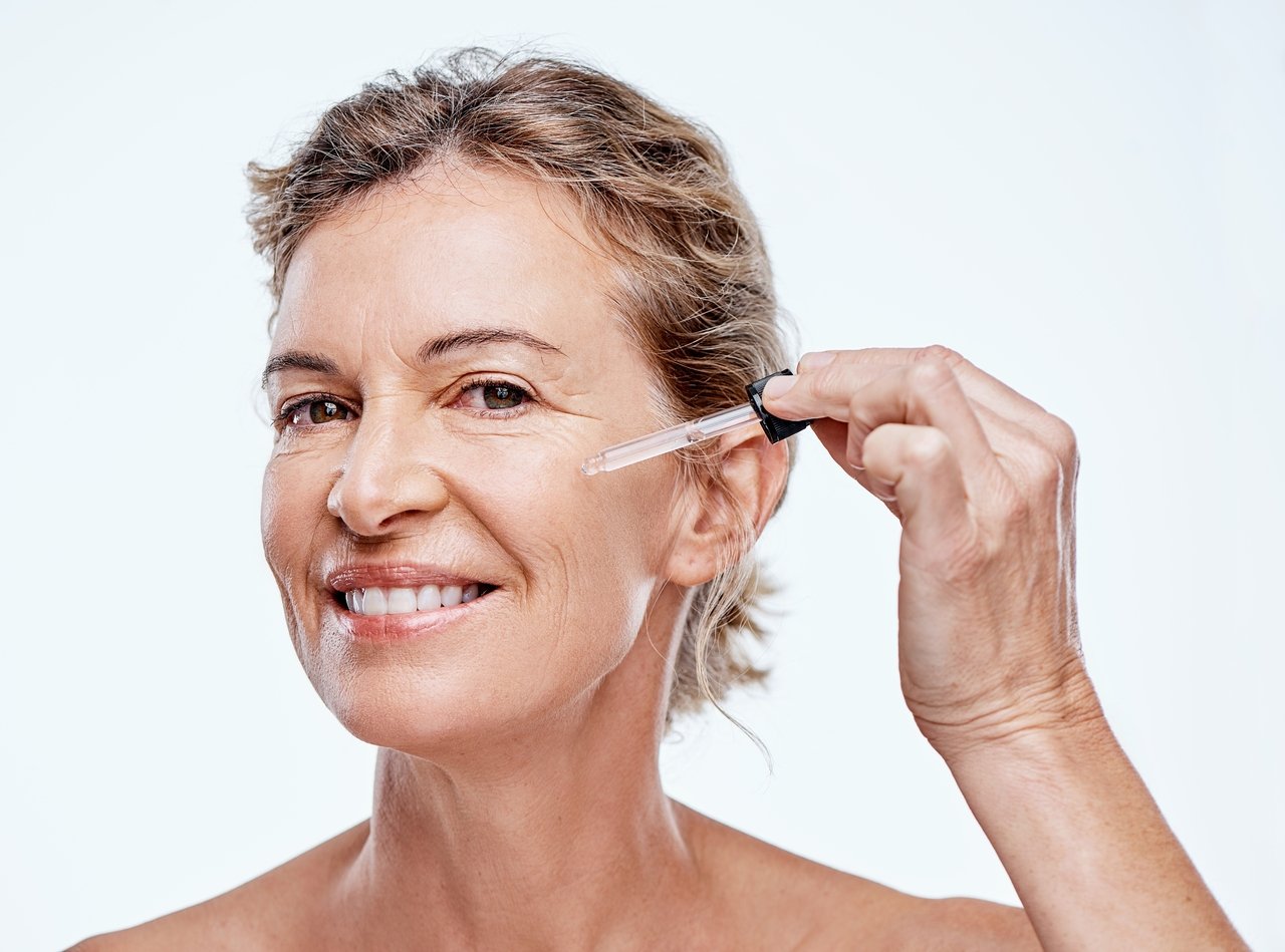 Foto de uma mulher sorrindo, aplicando um sérum com um conta-gotas no rosto, indicando tratamento com colágeno para flacidez.