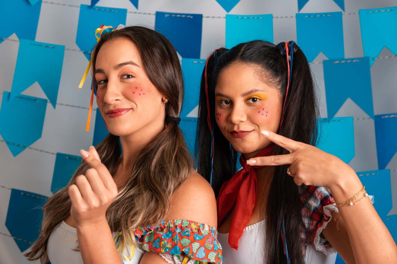 Duas mulheres sorrindo com maquiagem de festa junina e penteados temáticos, em frente a um fundo de bandeirinhas.